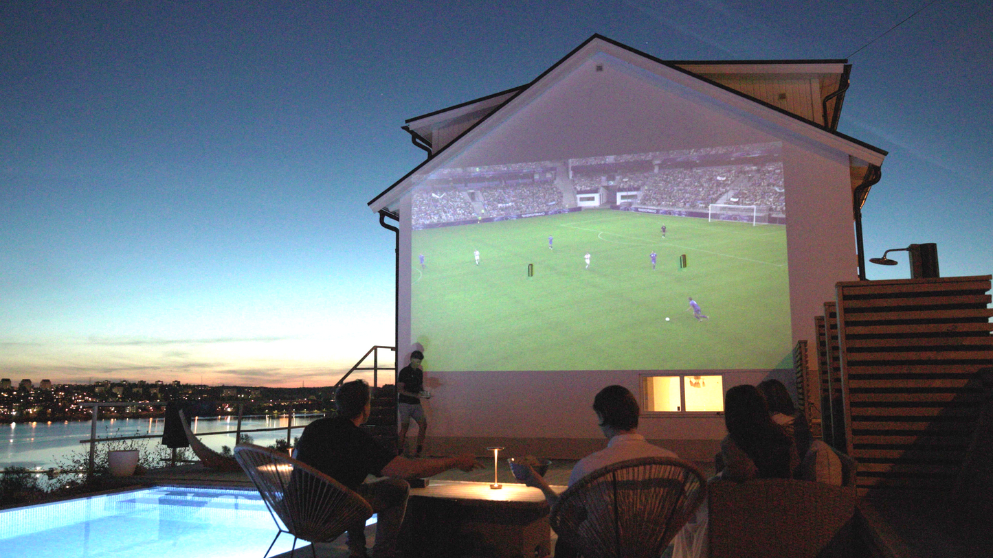 The world's smartest and portable projector, Estelle One, is placed on an outdoor table, projecting football onto an outdoor house wall. The picture is taken in the evening, and a group of people can be seen sitting around a table. One person is about to deliver drinks and is at the level of the projection. On the left side of the image, an illuminated pool is visible, and below it, there is a beautiful ocean view overlooking a bay. On the other side of the bay, lights from buildings can be seen.