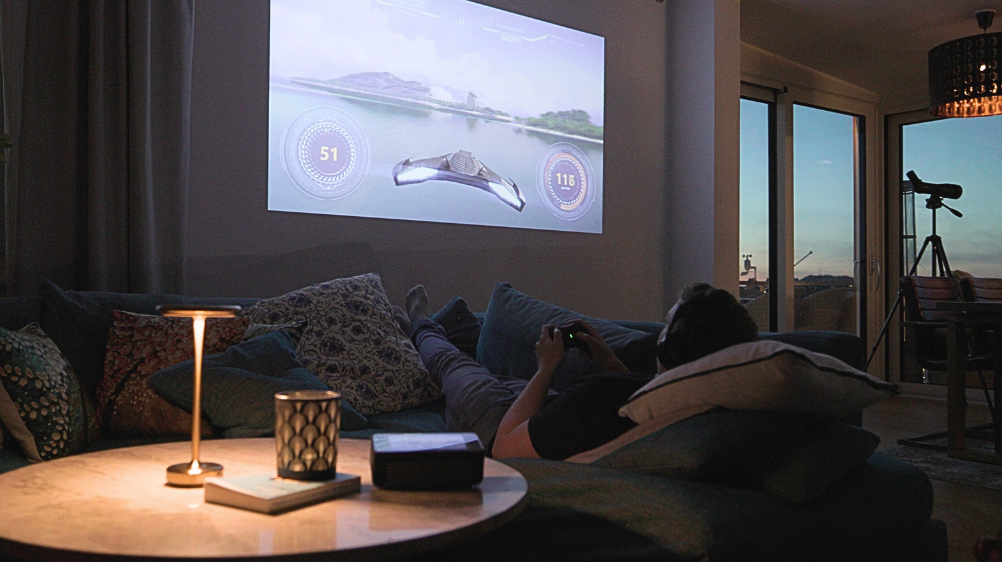The Estelle One projector sits on a round stone table, seen from behind. A lamp and a book with a light on it rest on the table. In front, a gray sofa with large cushions holds a person playing video games with headphones. The wall displays a tropical space scene projected by Estelle One. Outside, dusk is visible through the window, with binoculars nearby.