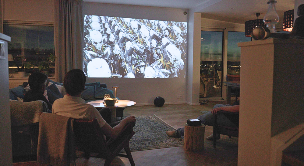 The Estelle One projector sits on a stump in the middle of a living room, displaying its touchscreen interface. The same image is projected large on the wall opposite. Three people are present: one on a sofa to the left, one in a chair to the left, and one in a chair to the right. On the glossy stone table in front of the sofa, there's a bowl of chips and a small lamp. It's evening outside, with distant residential lights visible.