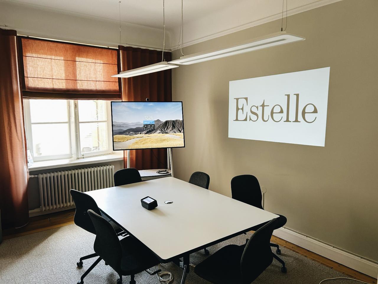 The world's smartest and portable projector, Estelle One, sits on a table in a meeting room. On the touchscreen, the Estelle logo is displayed in black against a white background, while the same image is projected in large format on the wall.