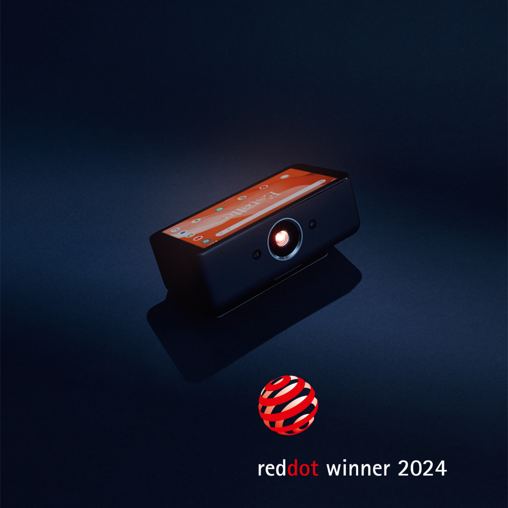 The Estelle One, the world's smartest and most portable projector, is featured in the center of the image with its touchscreen turned on and the projection lamp glowing. In the bottom right corner, there is a red and white Red Dot Winner 2024 logo. The background is a dark blue.