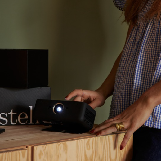 The world's smartest and most portable projector, Estelle One, is placed on a bench with the stand partially unfolded, revealing the projection lamp is on. A woman in a blue and white striped blouse navigates the touchscreen with her hands, adorned with rings and red nail polish. The bench is wood-colored. Behind the product on the bench stands the Estelle gift box, which is black with the Estelle logo printed in white. The wall behind is green.
