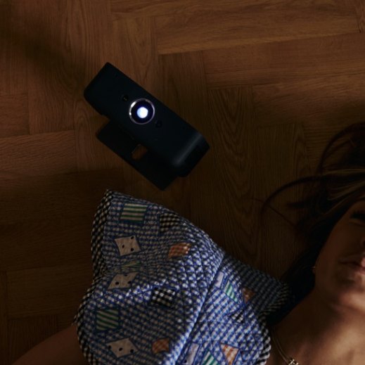 The world's smartest and most portable projector, Estelle One, is placed on a floor with the stand fully unfolded, projecting directly upwards. The image is captured from above, looking down towards the floor, showing the light cone and the fully unfolded stand. A woman lies beside Estelle One, looking towards the projection on the ceiling (not visible in the image), wearing a blue and white checkered blouse with square patterns in green, white, and red colors. The floor is made of oak and has an oak color.